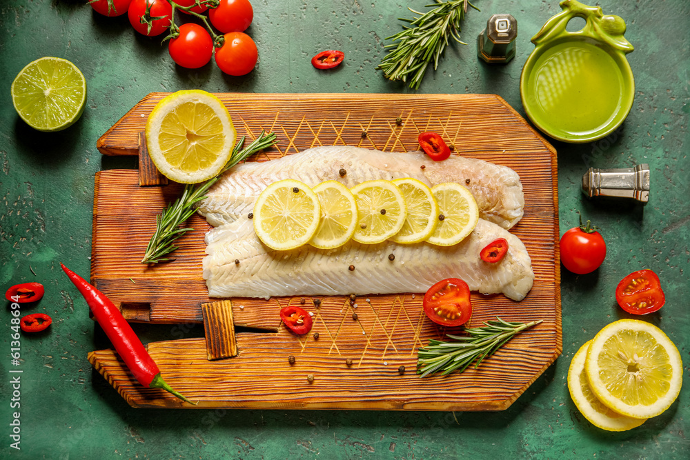 Wooden board with raw codfish fillet and ingredients on green background