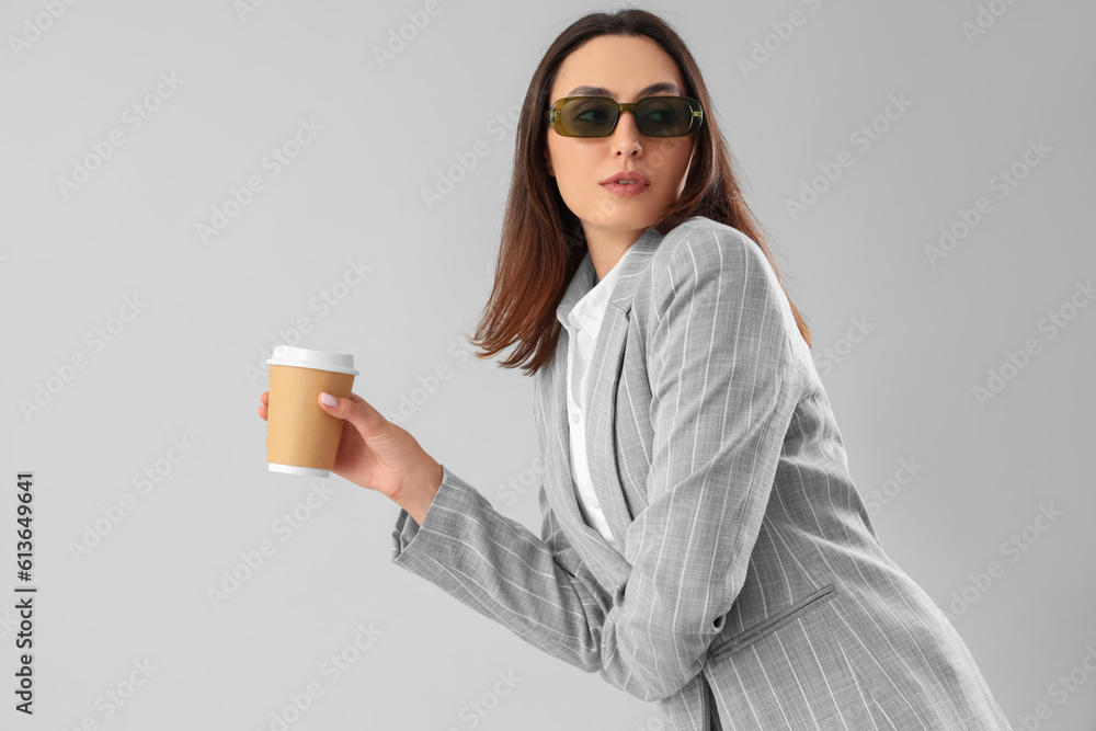 Young woman in stylish suit with cup of coffee on light background