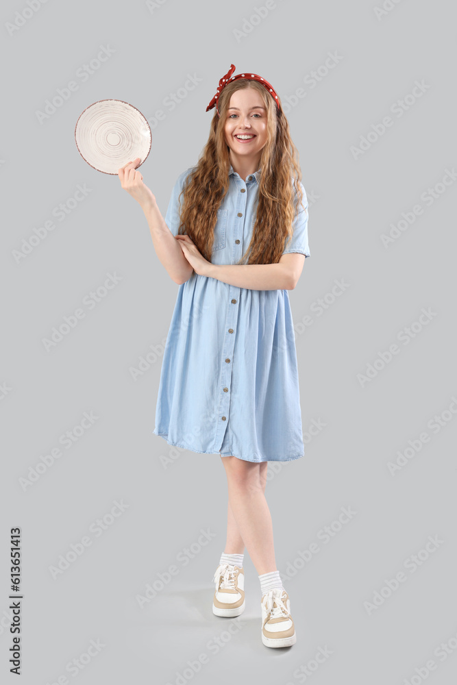 Beautiful young woman with empty plate on grey background