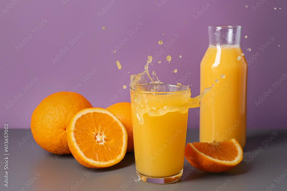 Bottle and glass of fresh orange juice with splashes on table against purple background