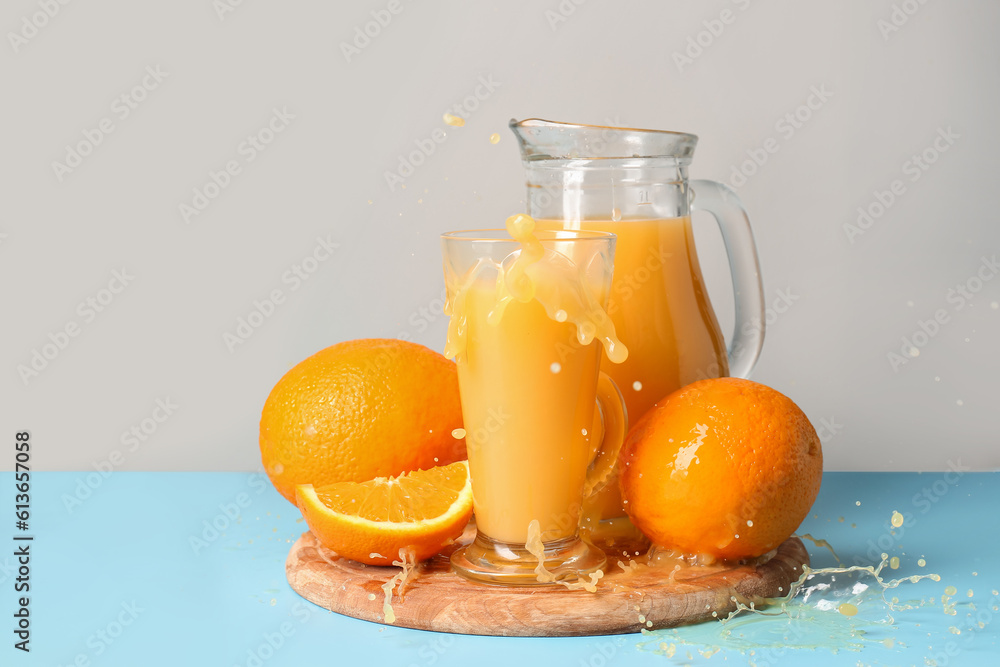 Jug and glass of fresh orange juice with splashes on table against grey background