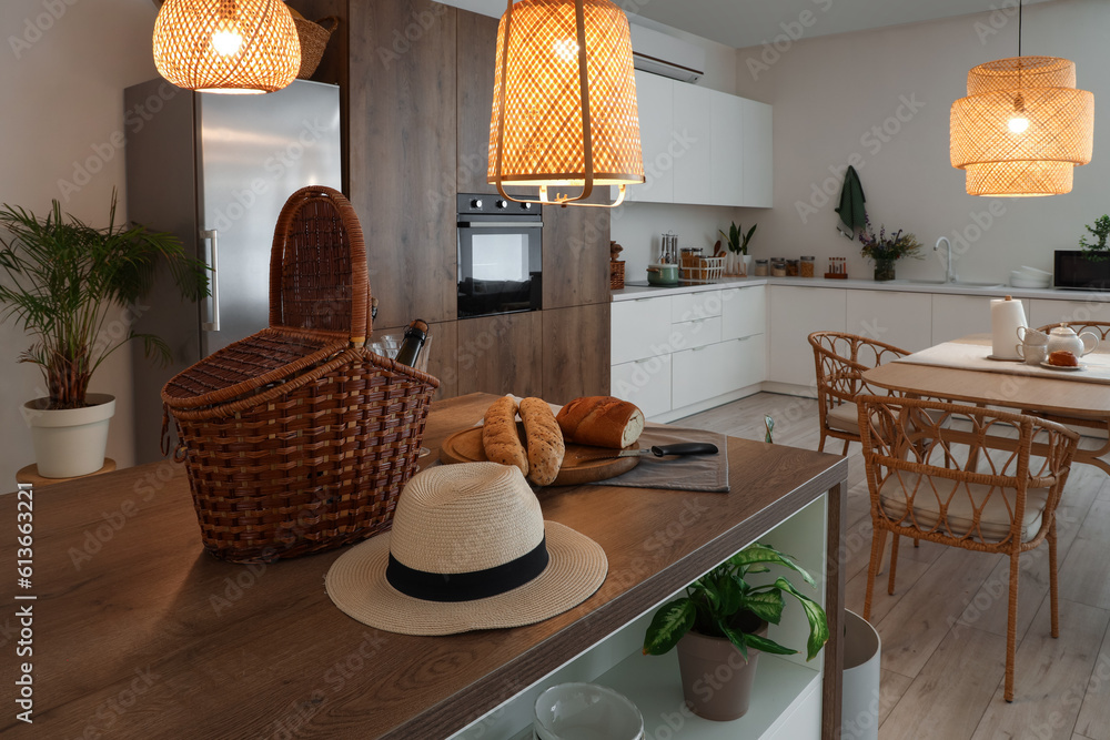 Hat, wicker basket and fresh bread on wooden island table in modern kitchen with glowing lamps