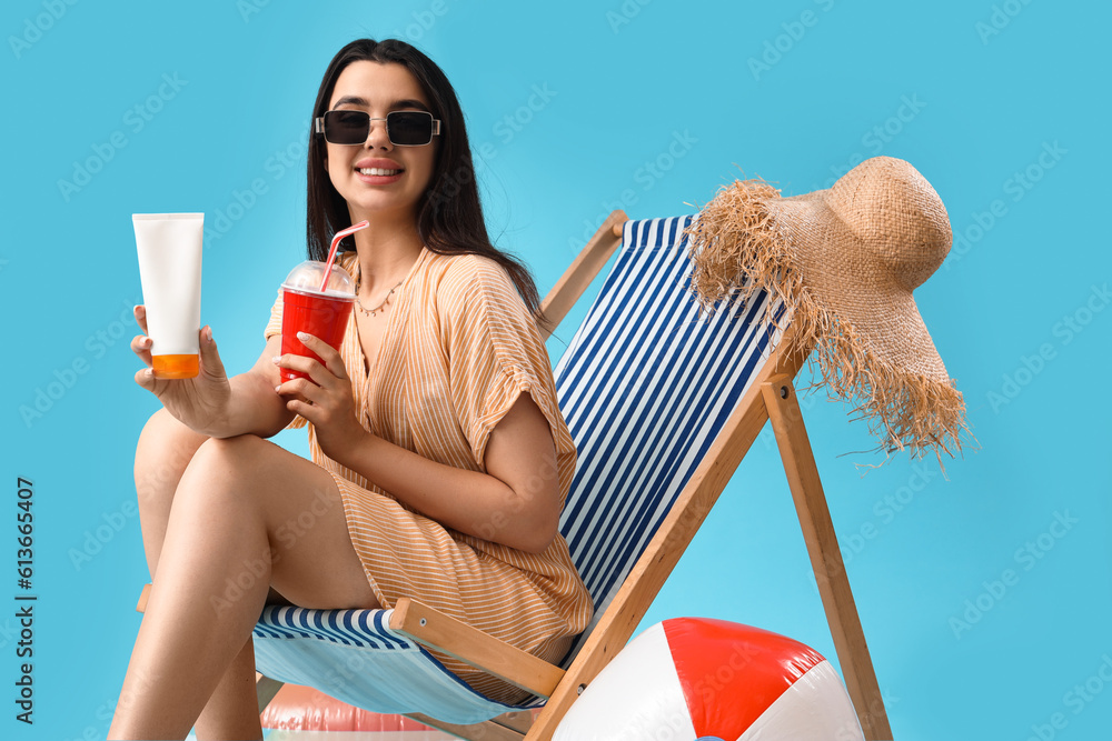 Young woman with sunscreen cream and soda in deck chair on blue background