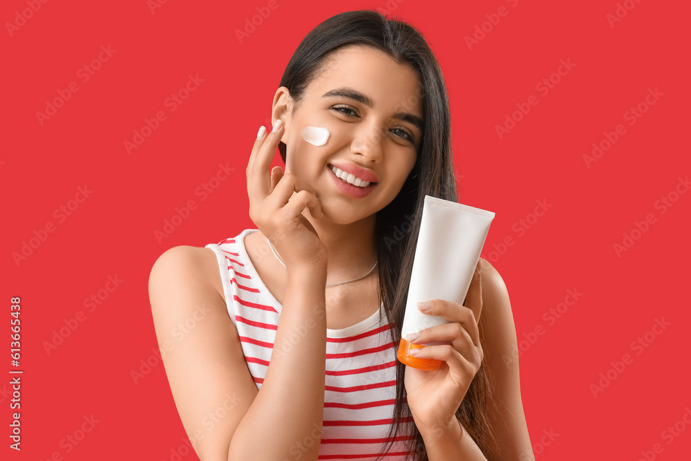 Young woman applying sunscreen cream onto her face against red background, closeup