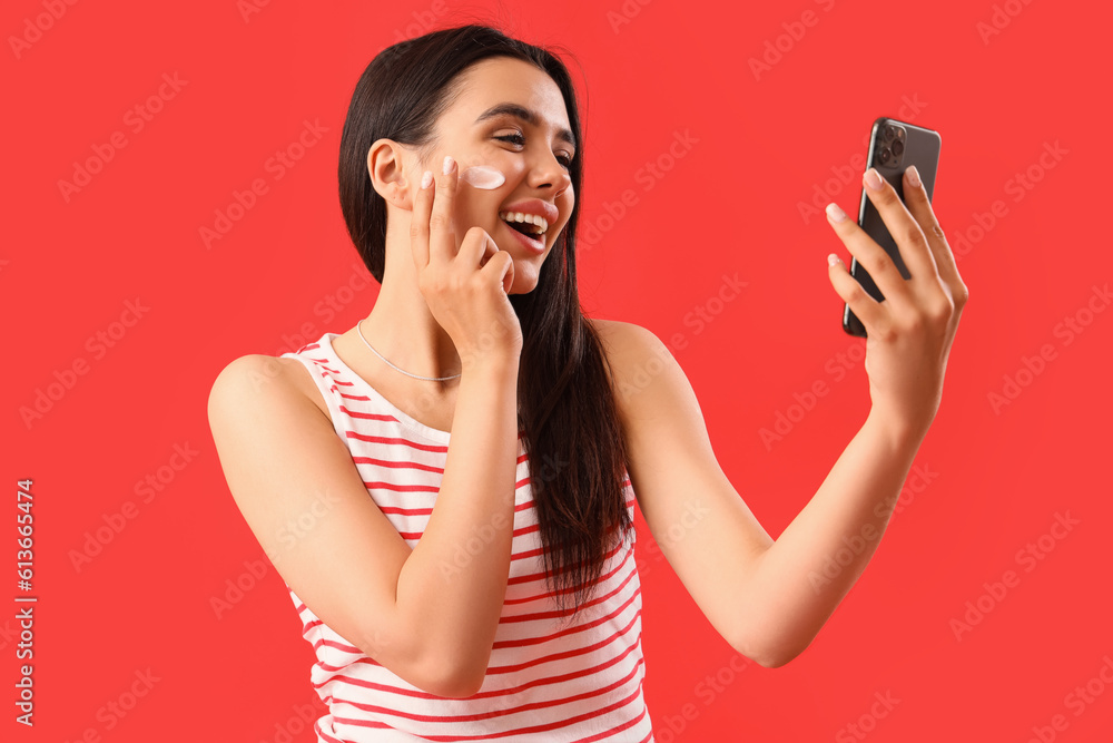 Young woman with mobile phone applying sunscreen cream onto her face against red background