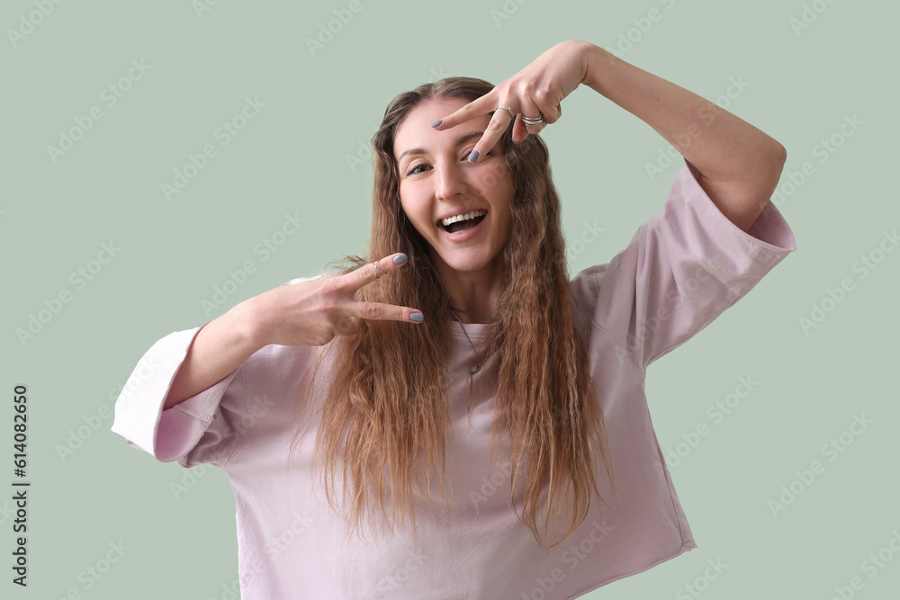 Beautiful young woman in pink sweatshirt showing victory gesture on green background