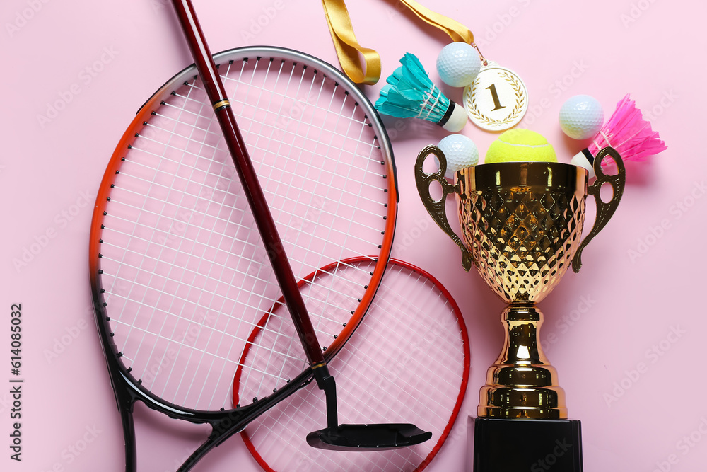 Gold cup with first place medal and sports equipment on pink background