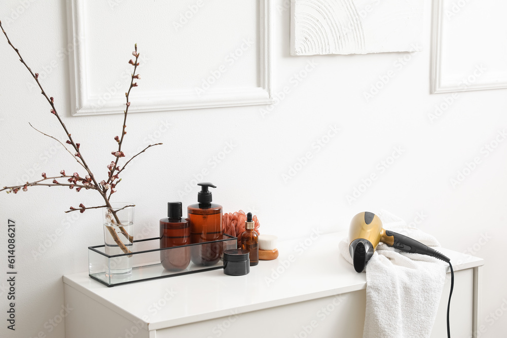 Bath accessories and vase with blooming tree branches on shelf near light wall