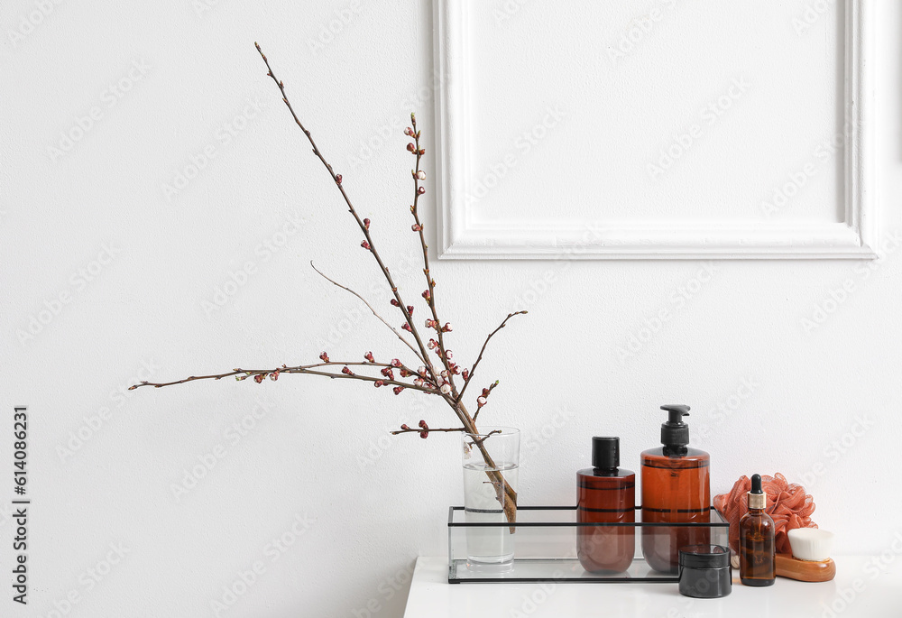 Bath accessories and vase with blooming tree branches on shelf near light wall