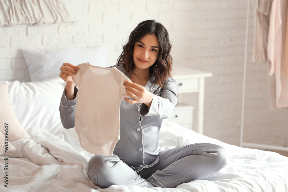 Young pregnant woman with baby bodysuit sitting in bedroom