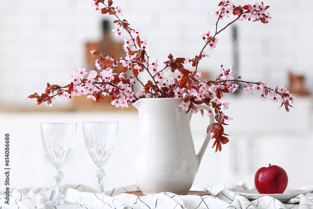 Vase with blooming tree branches, apple and glasses on table in kitchen