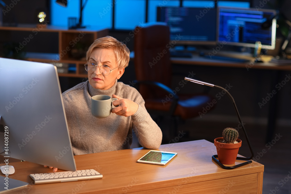 Mature female programmer with cup of coffee working in office at night