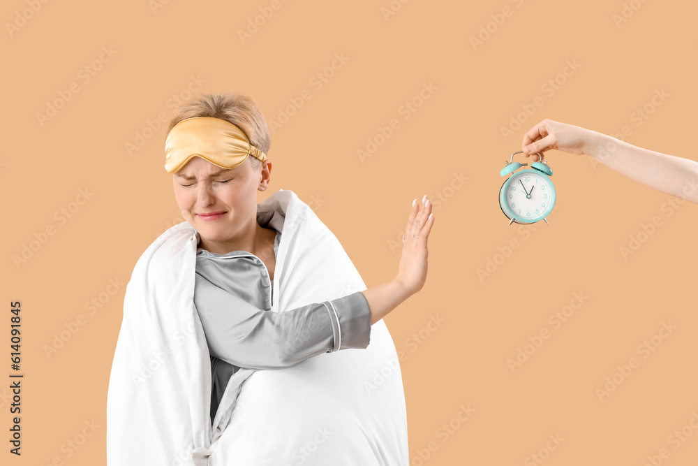 Upset young woman with sleeping mask and blanket rejecting alarm clock on beige background