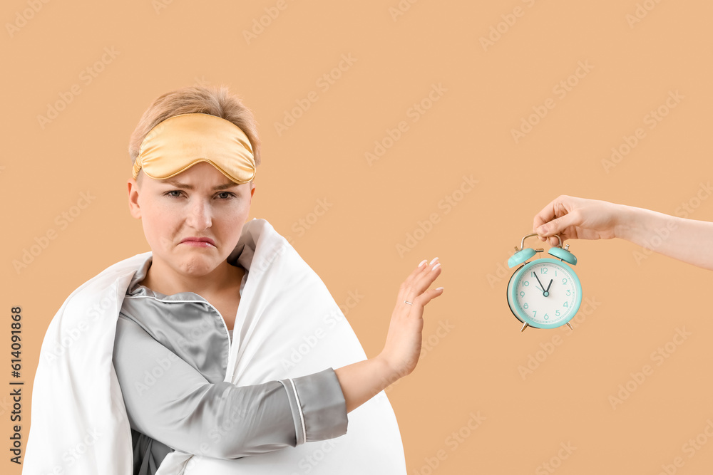 Displeased young woman with sleeping mask and blanket rejecting alarm clock on beige background