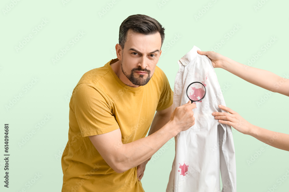 Handsome man with magnifier examining stained shirt on green background