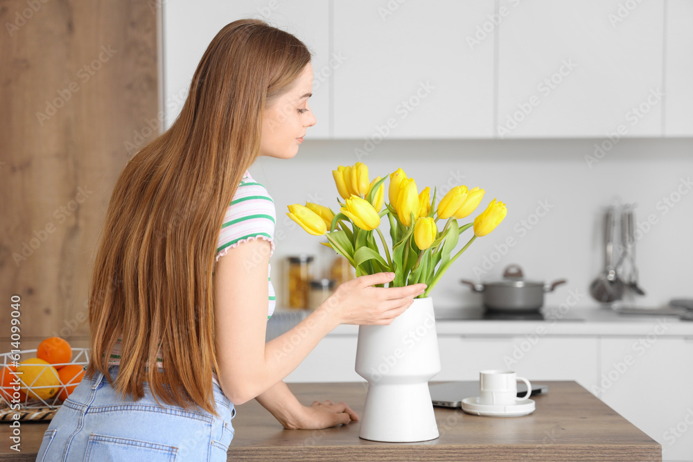 Beautiful young woman smelling blooming tulip flowers in modern kitchen