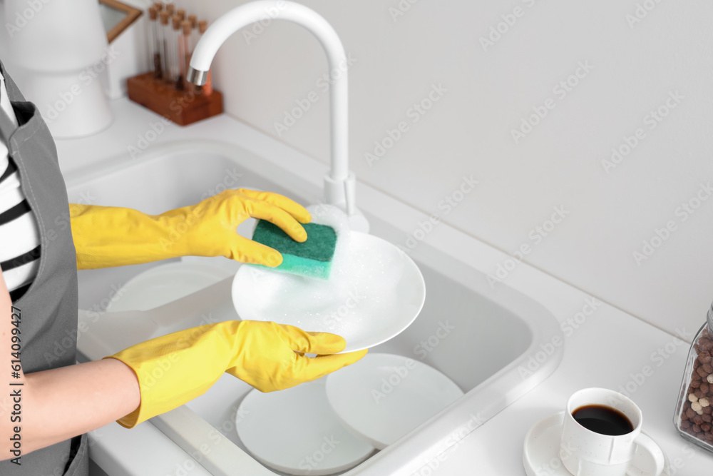 Woman in yellow rubber gloves washing plate with sponge in light kitchen