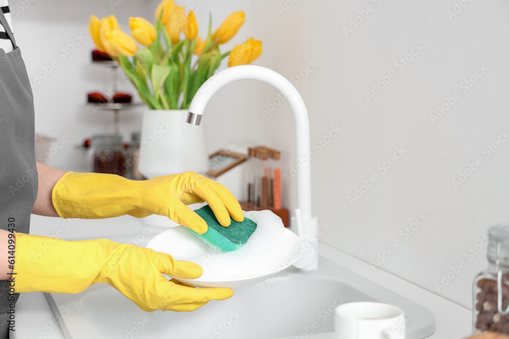 Woman in yellow rubber gloves washing plate with sponge in light kitchen