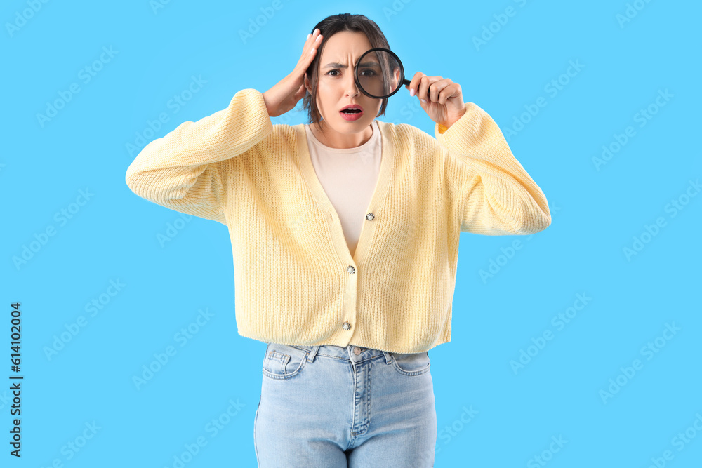 Shocked young woman with magnifier on blue background