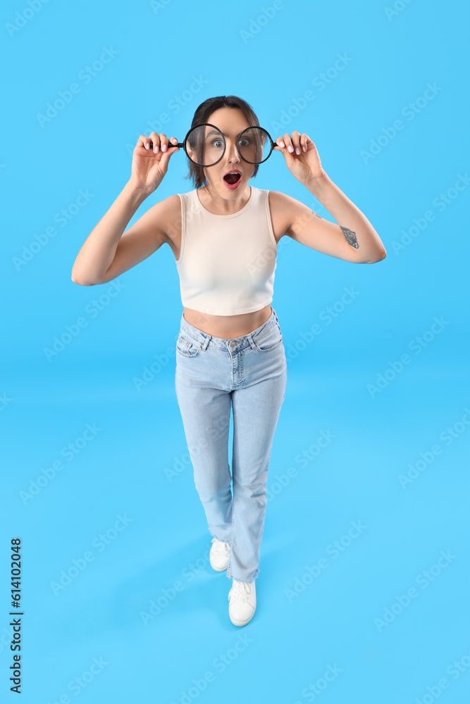 Surprised young woman with magnifiers on blue background