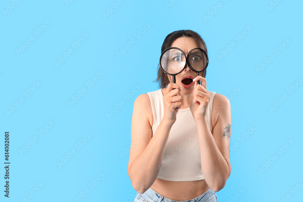 Shocked young woman with magnifiers on blue background