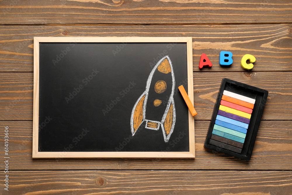 Chalkboard with drawn rocket, chalks and letters on brown wooden background