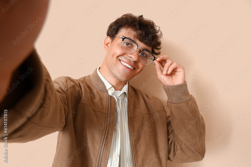 Young man in stylish eyeglasses taking selfie on beige background