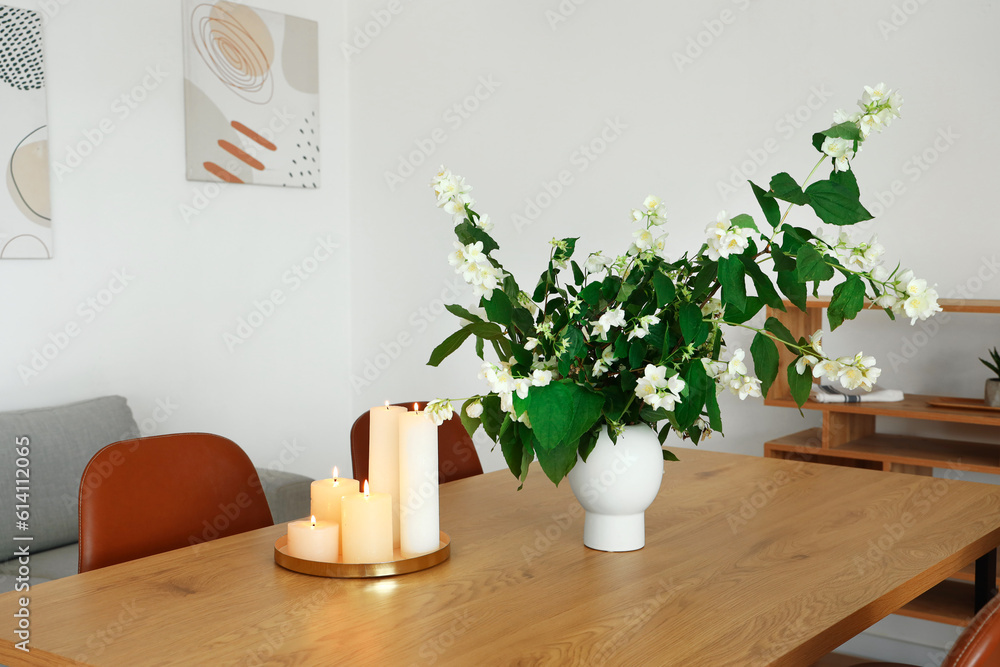 Vase with blooming jasmine flowers and burning candles on wooden table in modern kitchen