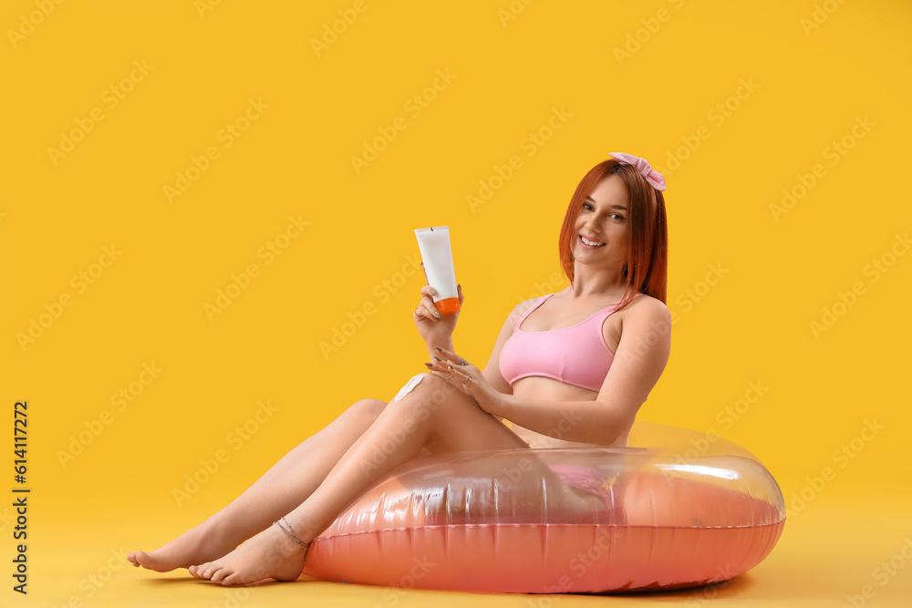 Young woman with inflatable ring applying sunscreen cream on yellow background