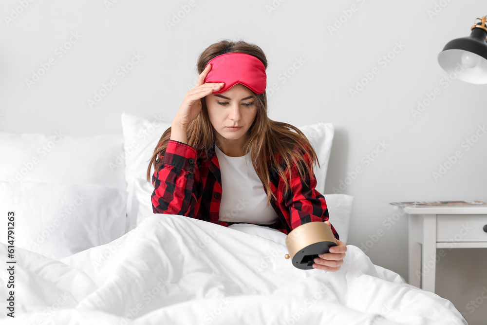 Upset young woman with alarm clock in bed at home