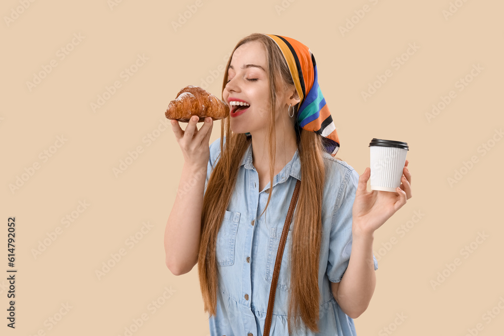 Young woman with cup of coffee eating croissant on beige background