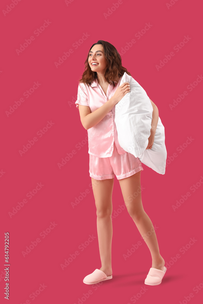 Young brunette woman in pajamas with pillow on pink background