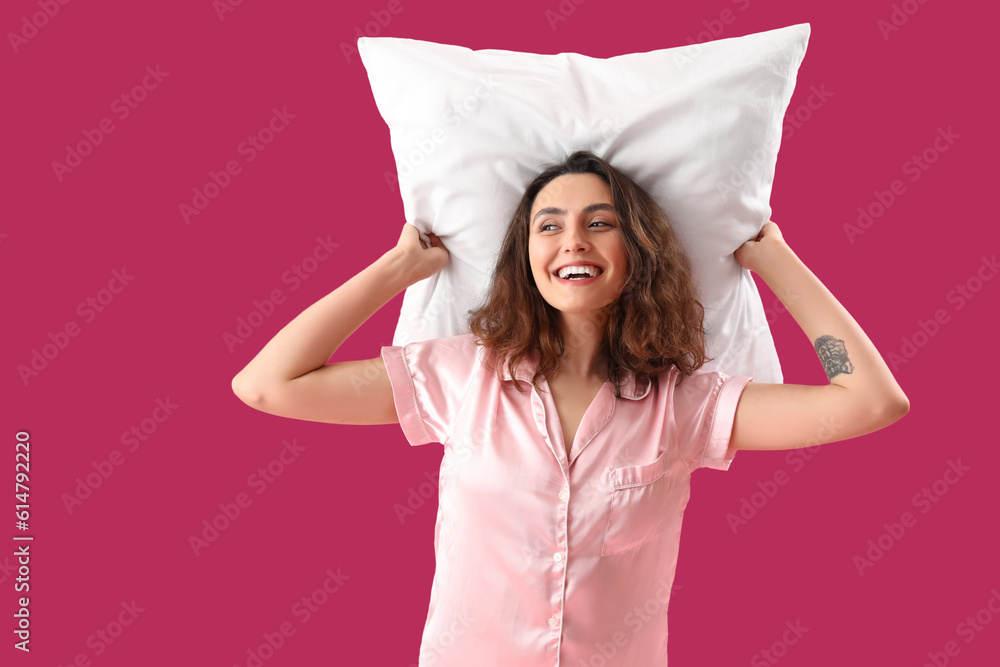 Young brunette woman in pajamas with pillow on pink background