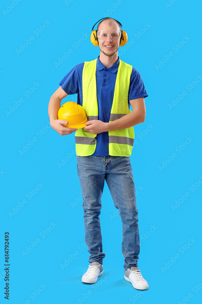 Male worker in vest, with hardhat and protective headphones on blue background