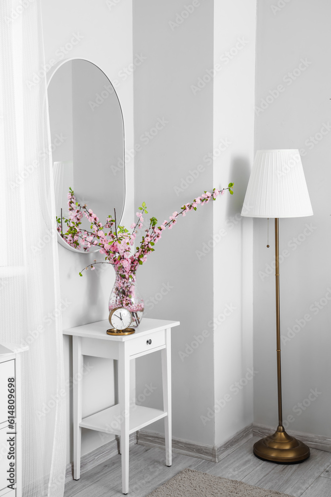 Vase with blooming sakura branches and alarm clock on end table in interior of light bedroom