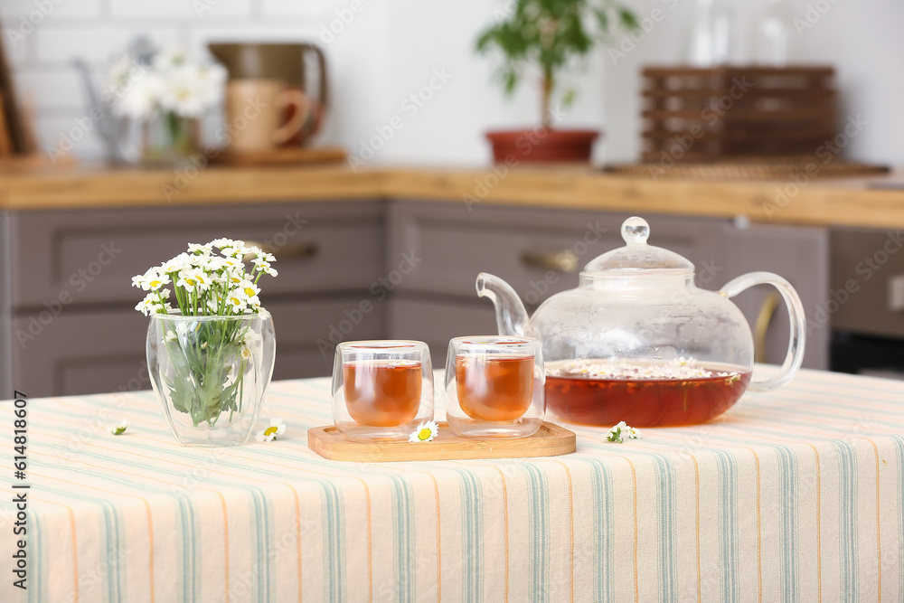 Wooden tray with teapot, cups of natural chamomile tea and flowers on table in kitchen