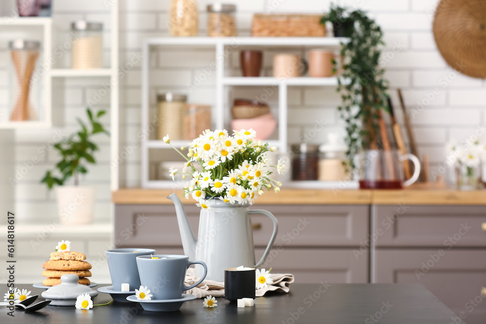 Watering can with flowers, cups of natural chamomile tea and cookies on black table in kitchen