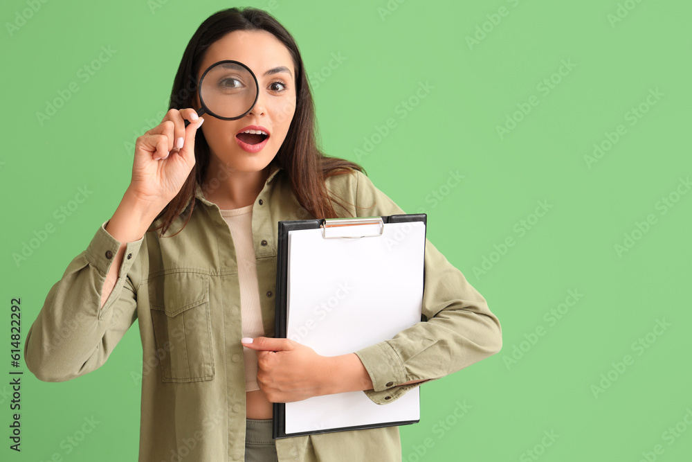 Surprised young woman with magnifier and clipboard on green background