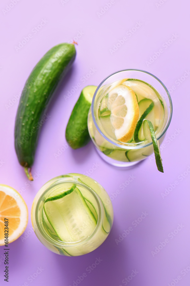 Glass and bottle of infused water with cucumber slices on lilac background