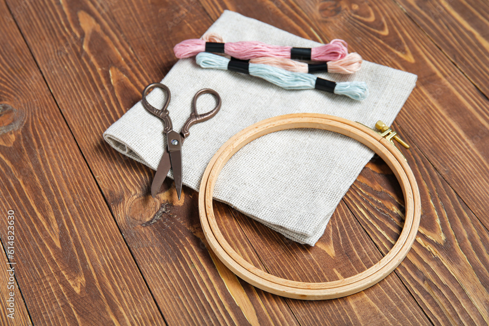 Wooden embroidery hoop with mouline threads, scissors and canvas on brown wooden background