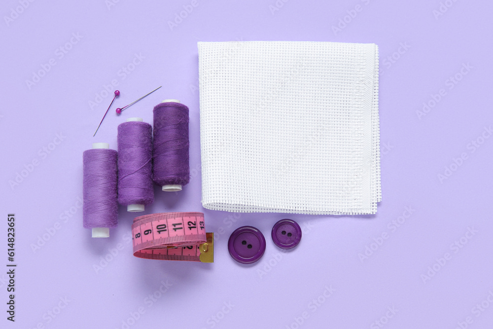 Canvas with buttons, thread spools and measuring tape on lilac background