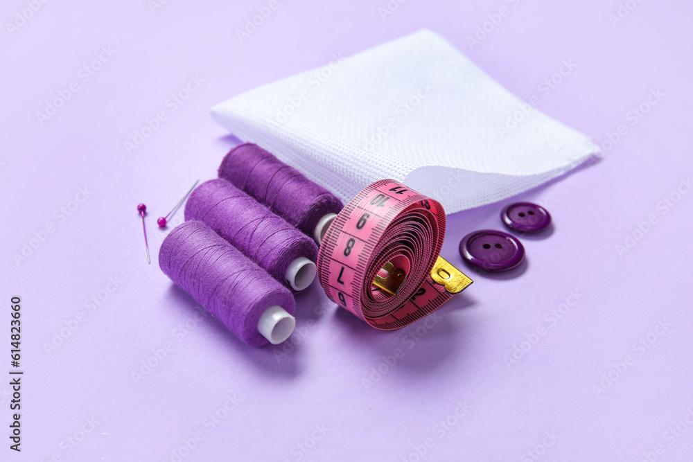 Canvas with buttons, thread spools and measuring tape on lilac background