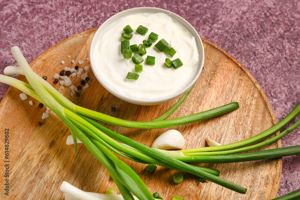 Board with bowl of tasty sour cream and sliced green onion on purple background, closeup