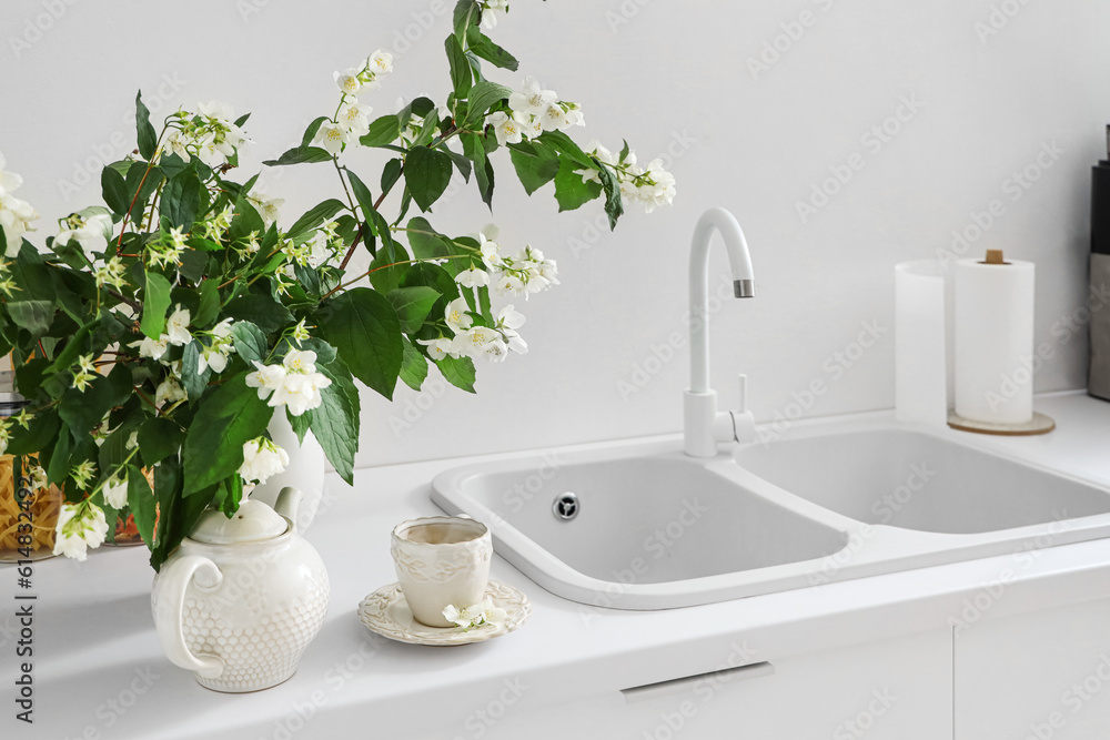 Vase with blooming jasmine flowers, teapot and cup of tea on white kitchen counter