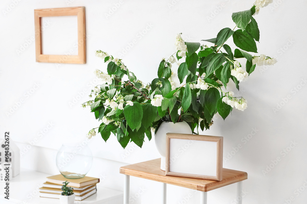 Vase with blooming jasmine flowers and blank frame on shelving unit in living room, closeup