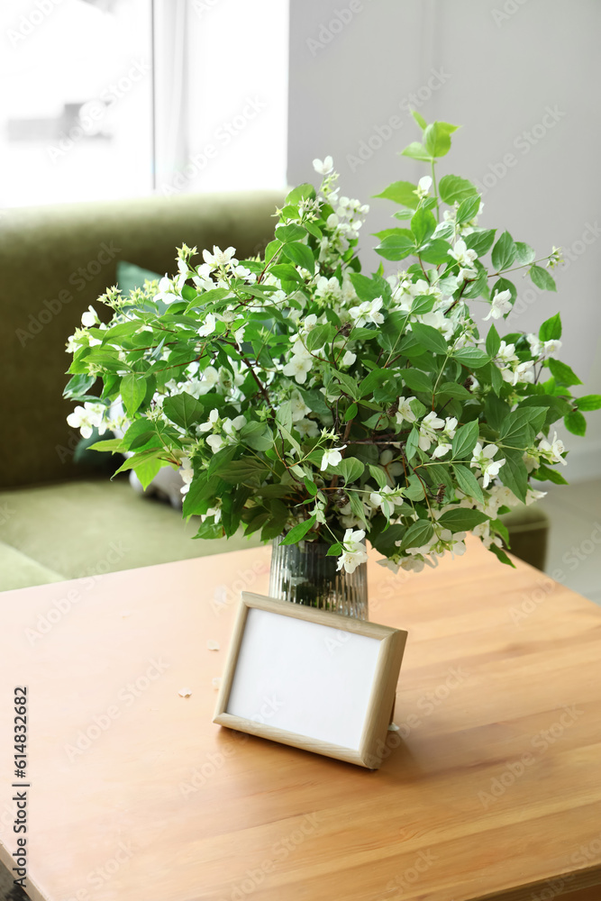 Vase with blooming jasmine flowers  and blank frame on wooden coffee table in living room