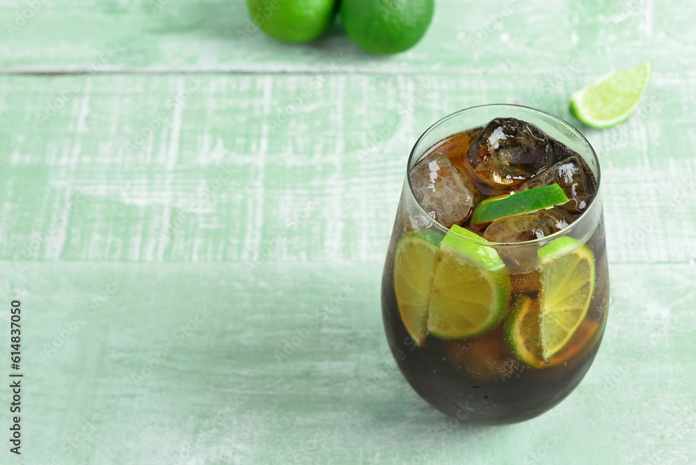 Glass of cold Cuba Libre cocktail and limes on green wooden table