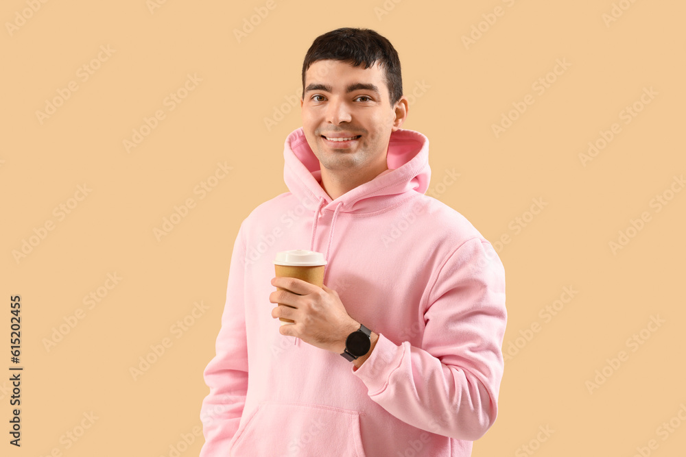 Stylish young man in hoodie drinking coffee on beige background