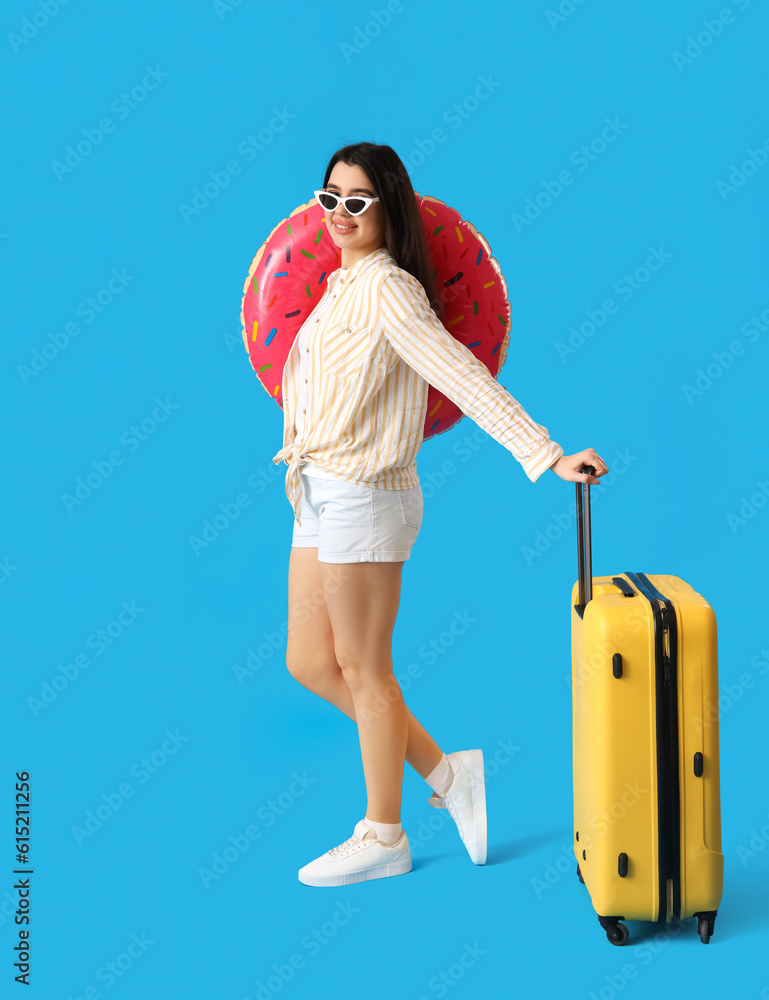 Young woman with inflatable ring and suitcase on blue background