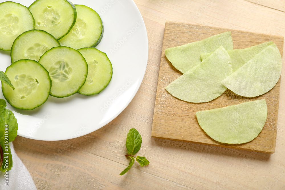 Board with cotton under-eye patches and cucumber slices on light wooden background, closeup